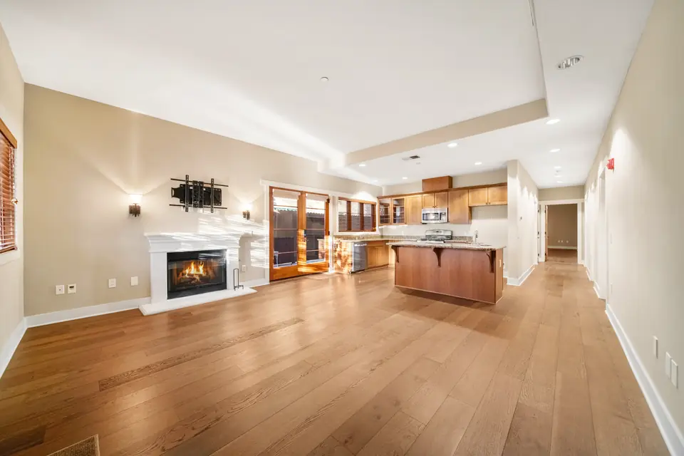 Living room with a fireplace and wood floors, kitchen beyond. Digitally added fire